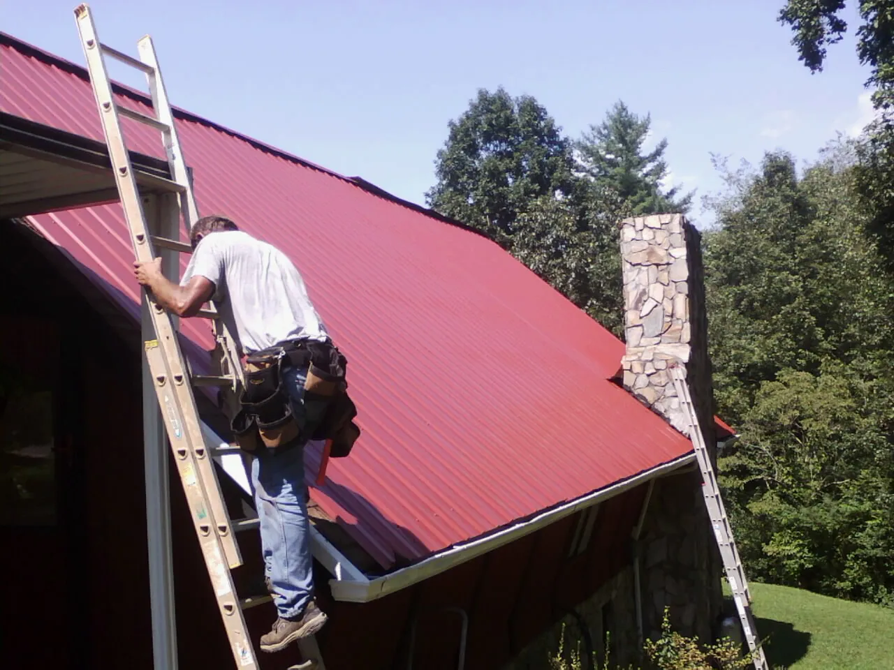 Roofer on a ladder beside a red metal roof and stone chimney — Soffit & Fascia Repair by Roofing Brothers in Mc Sherrystown